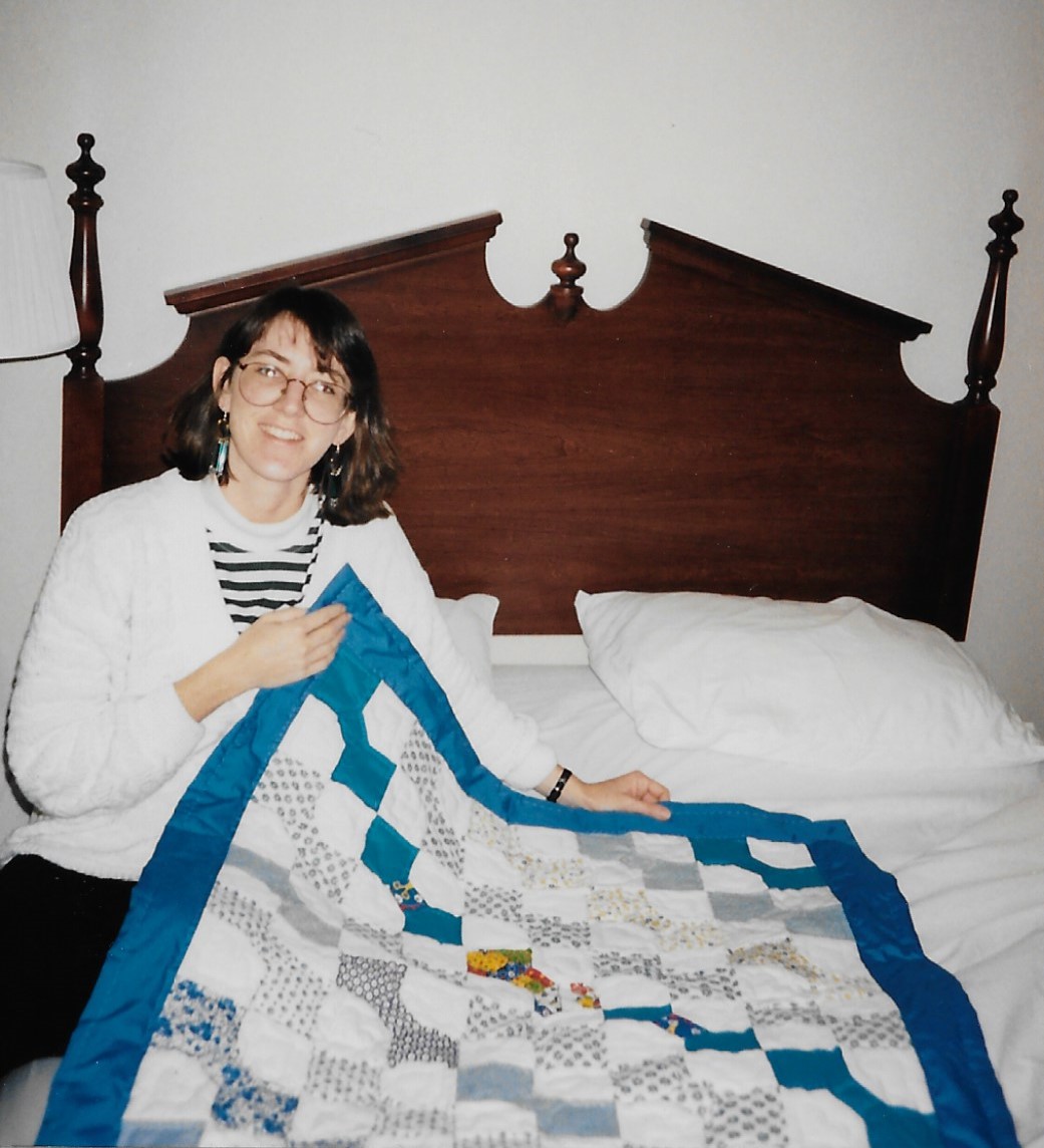 Sitting on bed holding up end of small bow tie quilt